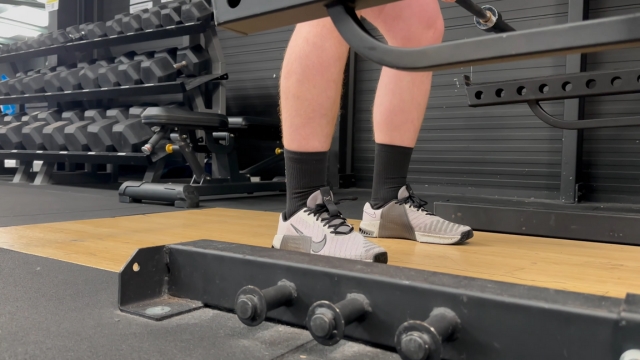 Muškarac u teretani nosi Nike Metcon 9 patike dok stoji ispod šipke za čučanj na platformi za dizanje tegova