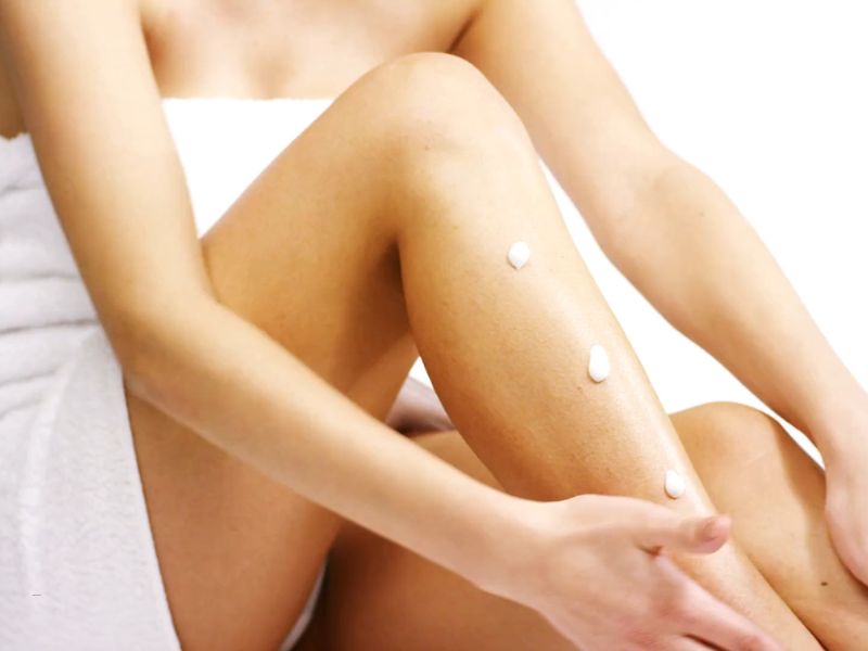 devojka sa peškirom oko tela sedi i maže mleko za telo na noge